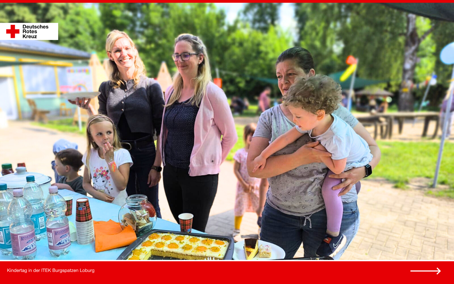 Eltern und Kinder stehen am Kuchenbuffet beim DRK-Kindertag in der Kita Burgspatzen Loburg; im Vordergrund sind Wasserflaschen, Kuchen und Becher zu sehen.