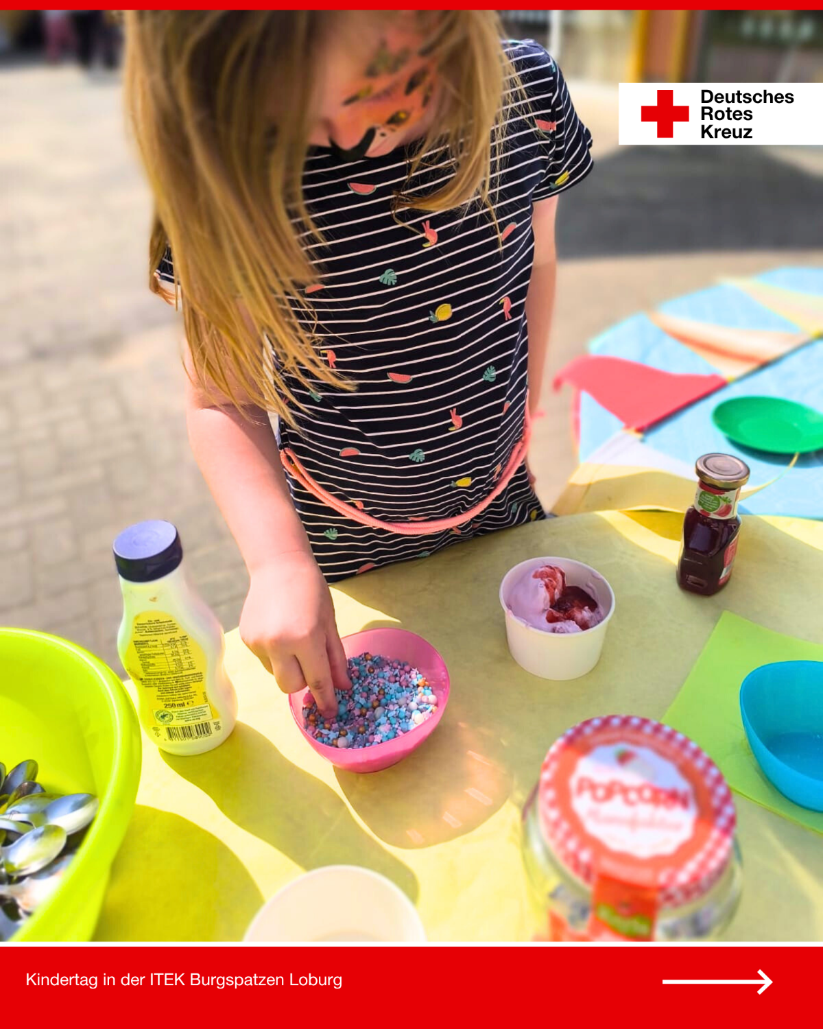 Ein Kind verziert seinen Eisbecher mit bunten Streuseln beim Kindertag in der DRK-Kita Burgspatzen Loburg; auf dem Tisch stehen weitere Zutaten bereit.