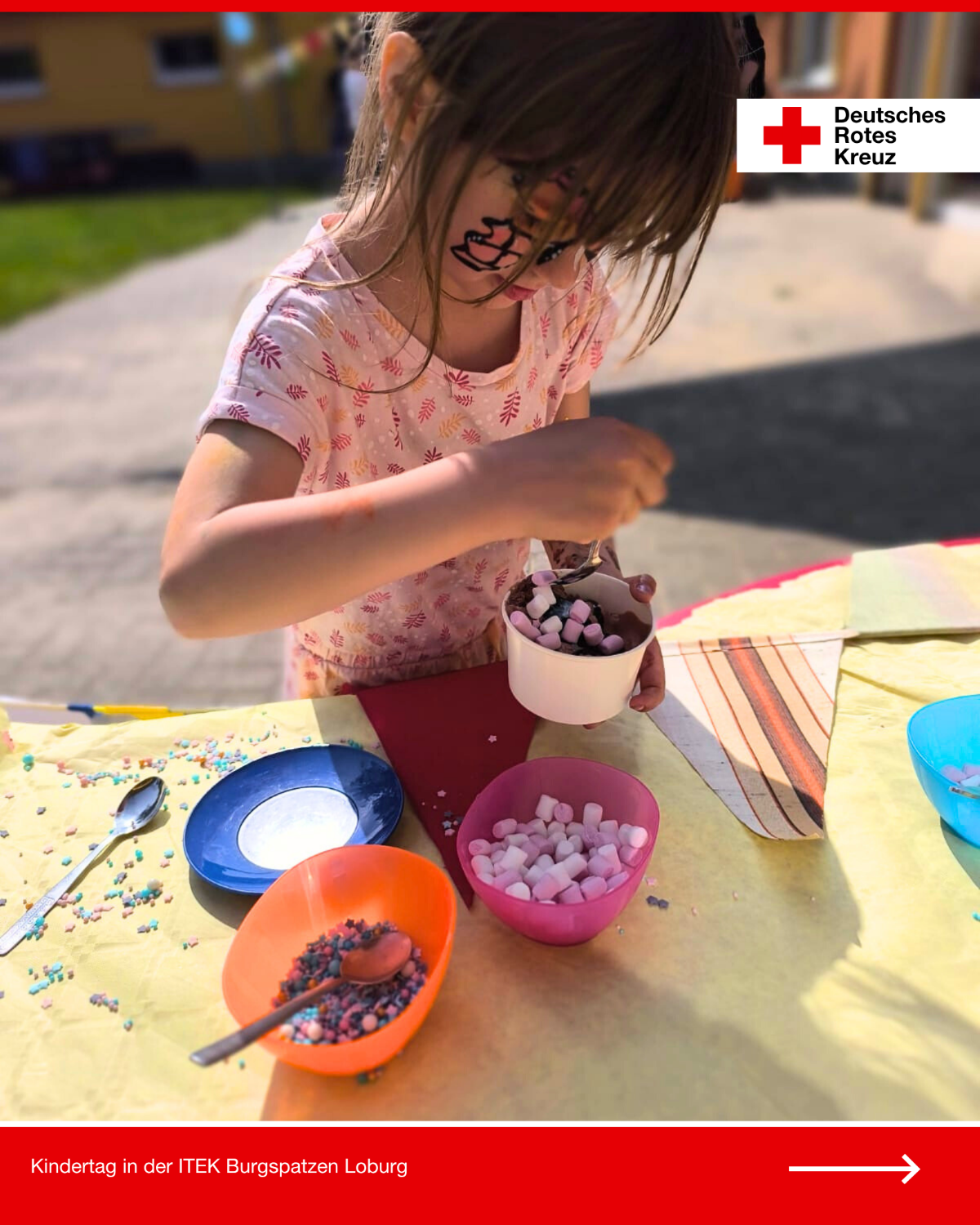Ein Kind mit bemaltem Gesicht befüllt beim Kindertag in der DRK-Kita Burgspatzen Loburg seinen Eisbecher mit Marshmallows und Streuseln.
