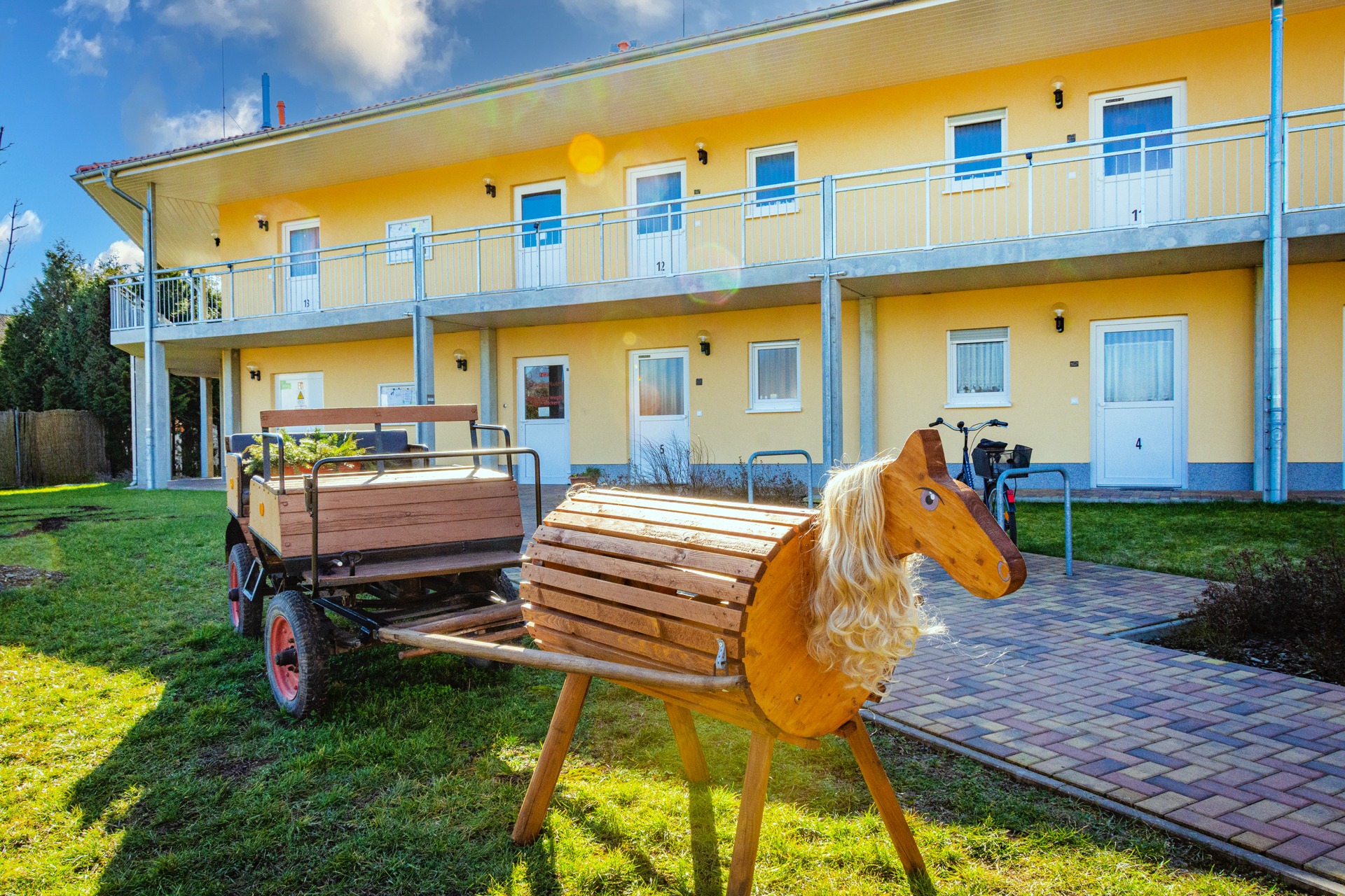 DRK-Tagespflege Möckern – Außenansicht Seniorenwohnpark Gelbes Gebäude der DRK-Tagespflege Möckern mit Balkon und Außenbereich, im Vordergrund ein Holzspielpferd mit Wagen.
