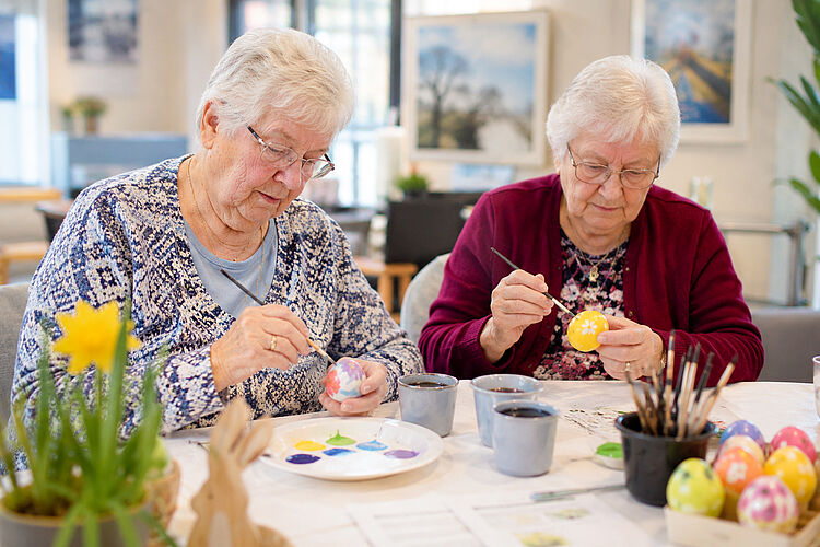 Zwei Seniorinnen bemalen gemeinsam bunte Ostereier im Rahmen eines kreativen Beschäftigungsangebotes in der Tagespflege.