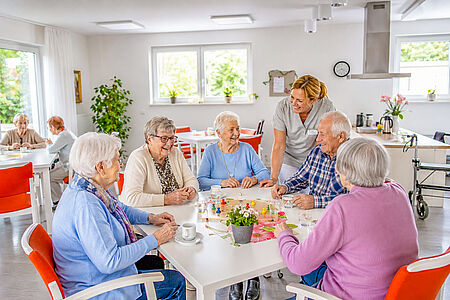 Seniorinnen und Senioren spielen gemeinsam mit einer Mitarbeiterin in einem hellen Gemeinschaftsraum der Tagespflege.