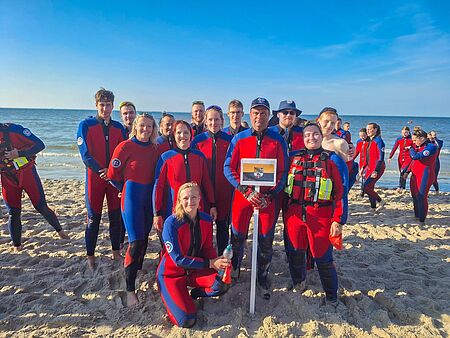 Gruppenfoto von jungen DRK-Wasserwacht-Mitgliedern im Neoprenanzug am Ostseestrand; sie stehen lächelnd im Sand, im Vordergrund wird ein Schild mit der Flagge Sachsen-Anhalts und dem DRK-Wasserwacht-Logo gehalten.