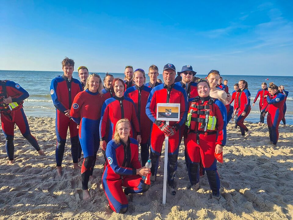 Gruppenfoto von jungen DRK-Wasserwacht-Mitgliedern im Neoprenanzug am Ostseestrand; sie stehen lächelnd im Sand, im Vordergrund wird ein Schild mit der Flagge Sachsen-Anhalts und dem DRK-Wasserwacht-Logo gehalten.