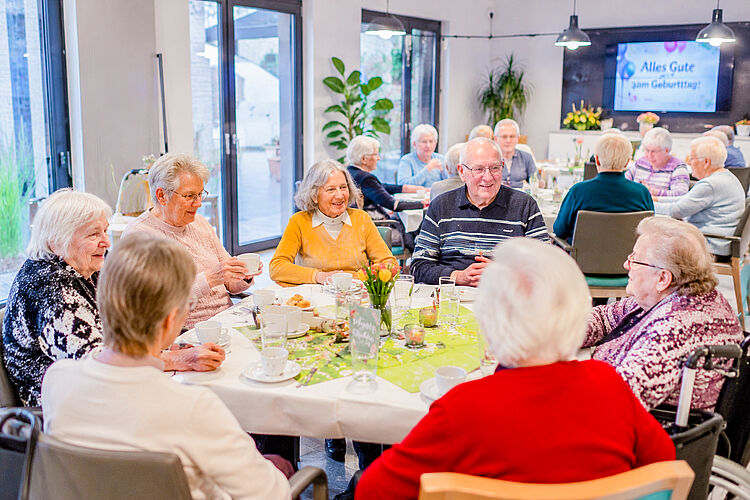 Seniorinnen und Senioren sitzen bei einer gemeinsamen Geburtstagsfeier in der Tagespflege an festlich gedeckten Tischen.