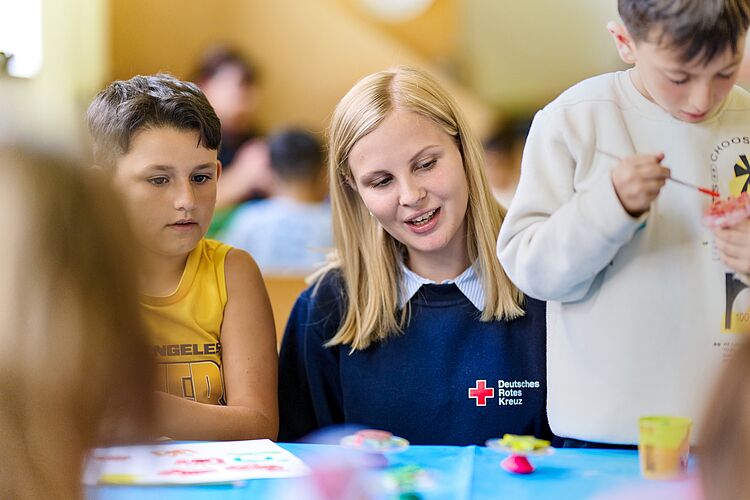 Eine DRK-Mitarbeiterin sitzt mit mehreren Kindern an einem Tisch und begleitet sie beim Basteln und Malen. Die Szene zeigt konzentrierte und fröhliche Kinder in einer kreativen Hort-Umgebung.