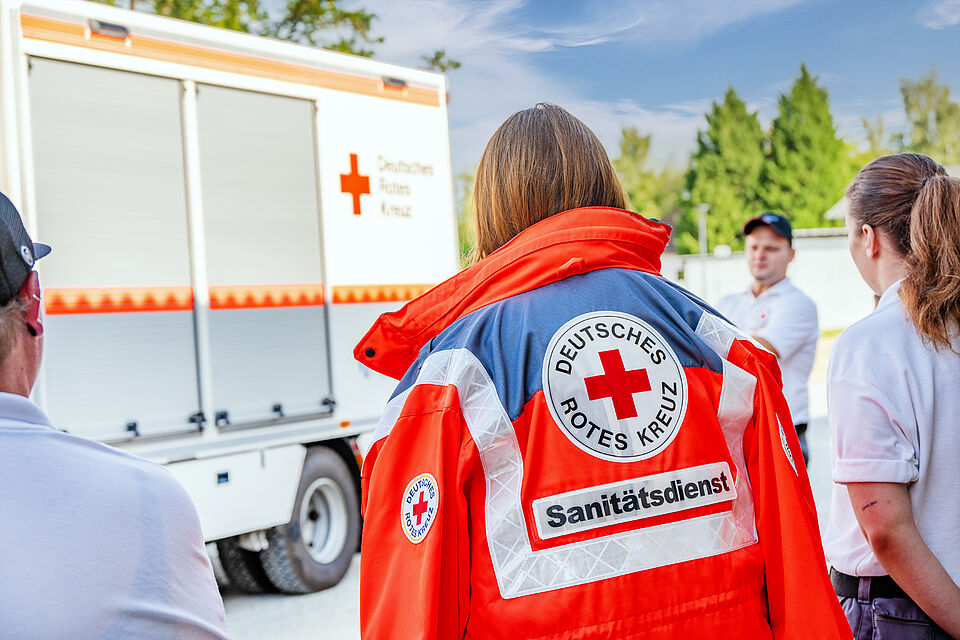 DRK-Sanitäterin in Einsatzjacke beim Dienstabend, während der Gruppenführer der Sanitätsgruppe den Ablauf zum Thema GW-San erklärt.