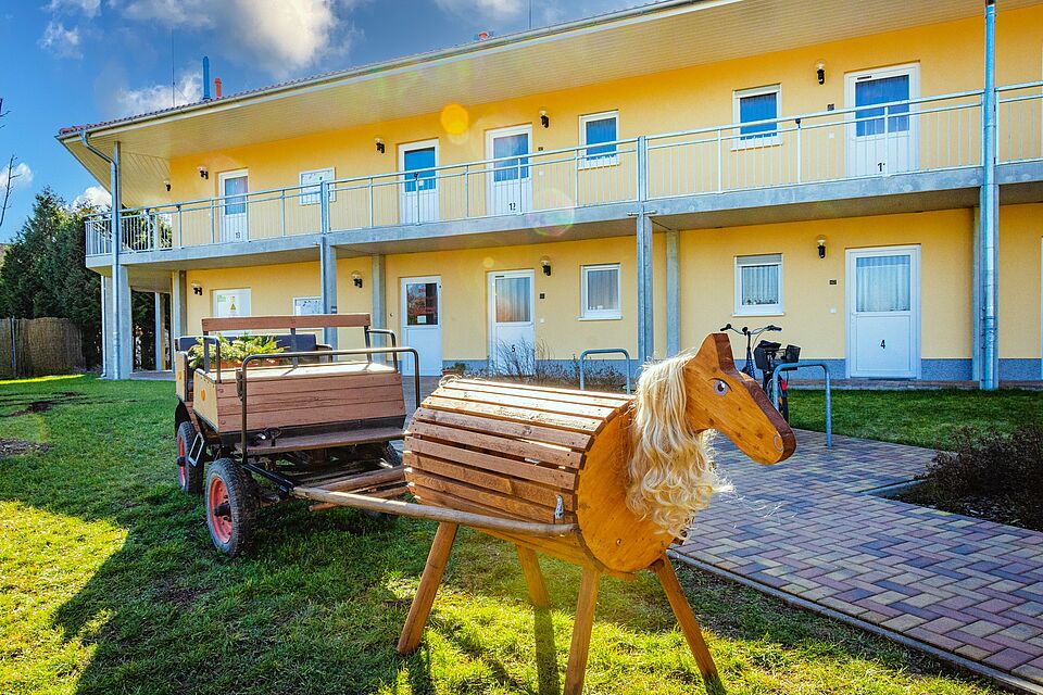 Gelbes Gebäude der DRK-Tagespflege Möckern mit Balkon und Außenbereich, im Vordergrund ein Holzspielpferd mit Wagen.