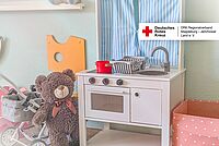Kinderküche mit blauem Vorhang, Spielzeug-Teddy und Kinderwagen als Titelmotiv der DRK-Spendenaktion.