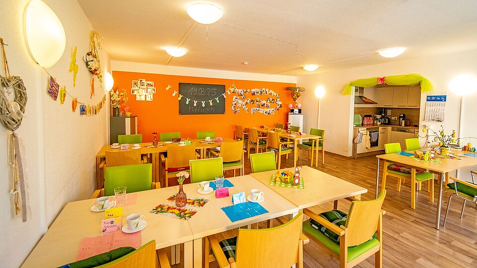 Innenaufnahme des bunt dekorierten Ess- und Aufenthaltsbereichs der DRK-Tagespflege Gommern mit Holztischen, grünen Stühlen, Blumen und Küchennische.
