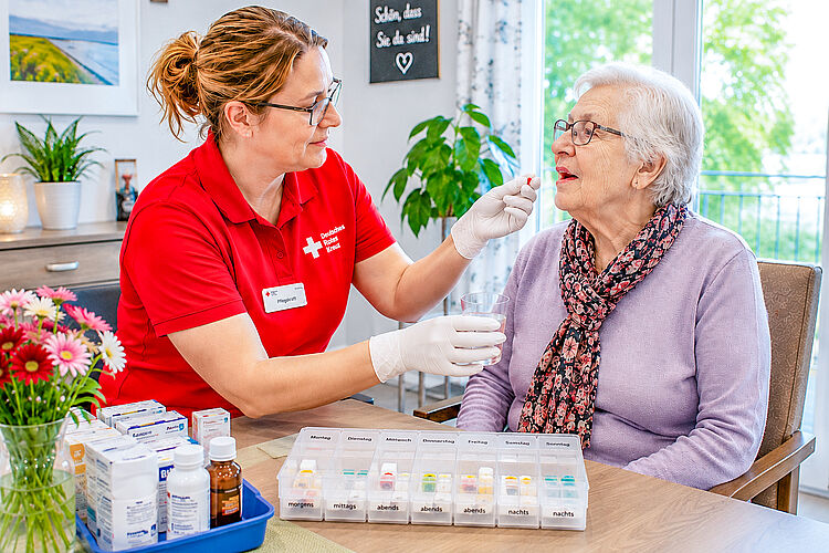 Eine Pflegekraft reicht einer Seniorin ein Medikament und hält ihr ein Glas Wasser bereit.
