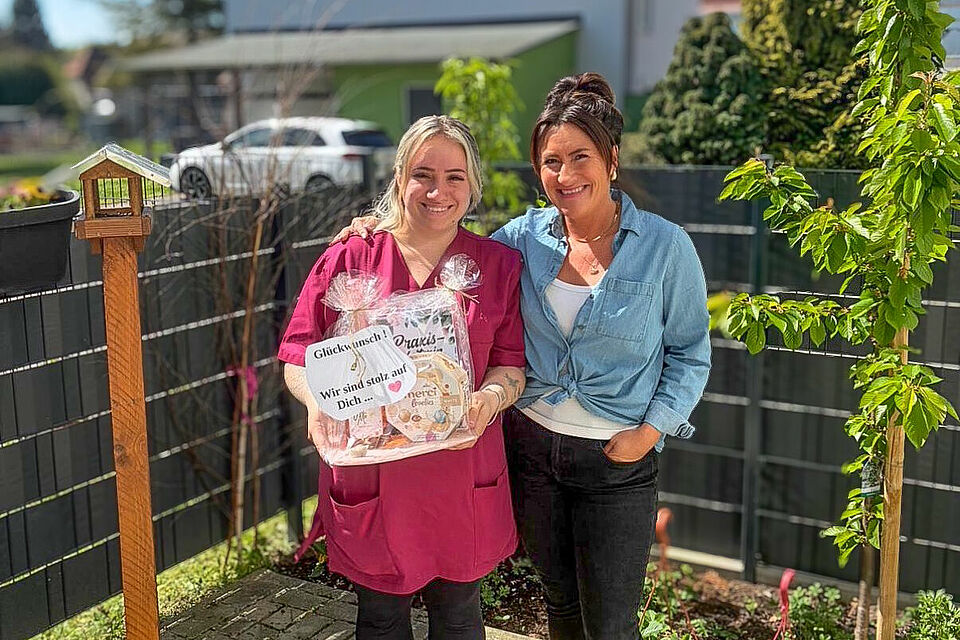 Zwei Frauen stehen im Außenbereich der DRK-Tagespflege Loburg; eine überreicht ein Geschenk zur bestandenen PAL-Weiterbildung