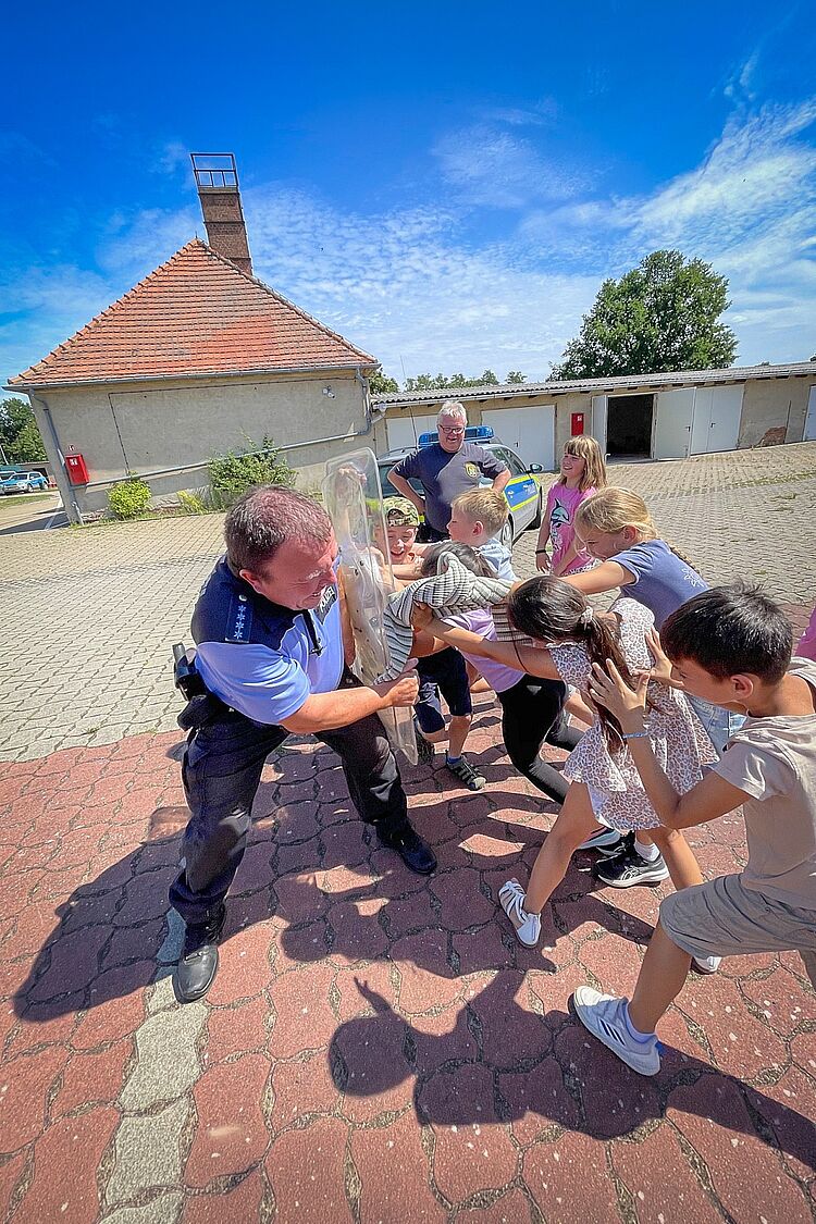 Hortkinder lernen die Schutzausrüstung der Polizei kennen Kinder des DRK-Hortes „Ludwig Uhland“ probieren gemeinsam mit einem Polizeibeamten ein transparentes Einsatzschild aus.