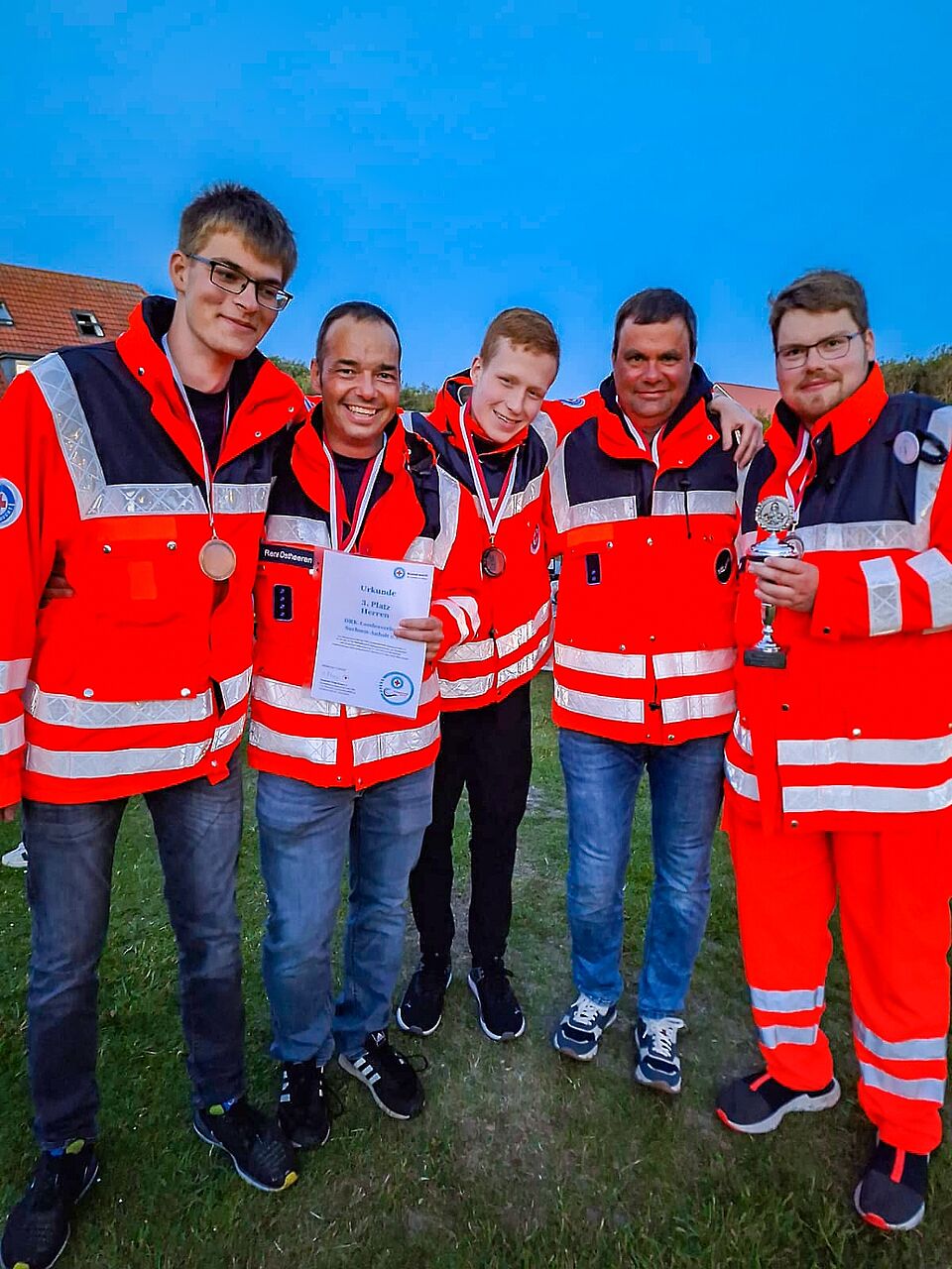 Fünf Mitglieder der DRK-Wasserwacht in roter Einsatzkleidung stehen eng beieinander, tragen Medaillen und halten stolz Urkunde und Pokal in die Kamera.